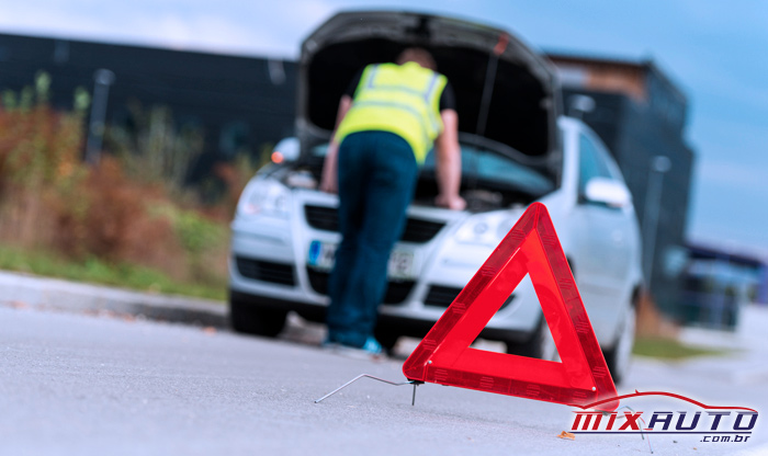 Triângulo para sinalização em foco e à frente de homem com colete de trânsito parado em frente a carro prateado com o capô levantado